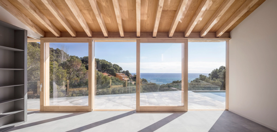 Interior of a detached single-family home with elegant contemporary architecture defined by double-height spaces, sloped ceilings with natural wood finishes, and large panoramic openings facing the landscape and the sea. Natural light flows throughout the space and reinforces the sense of spaciousness, while the combination of neutral materials and warm textures creates a serene, bright atmosphere fully connected to the outdoors.
