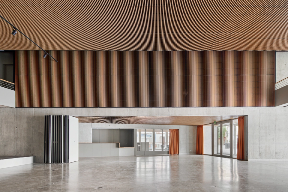 Main hall of a contemporary cultural center prepared to host performances, conferences, exhibitions, and community activities in a flexible and multipurpose space. The interior architecture stands out for its acoustic wooden slat ceilings, combined with exposed concrete and an open layout that promotes functionality and user circulation. The integrated stage and upper galleries reinforce the versatility of the cultural center, creating a modern, warm environment adapted to different cultural and social formats.