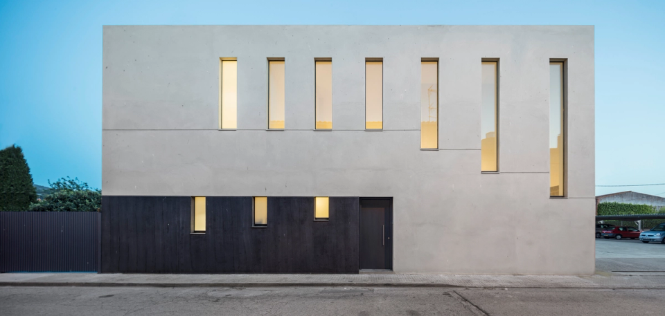 The main façade is presented as a compact and bold volume defined by pure lines and a minimalist composition of vertical openings that provide rhythm and depth. The contrast between the light finishes of the upper floor and the dark tones of the base reinforces the horizontal reading of the building, while interior lighting emphasizes the warmth and serene character of the home at dusk.