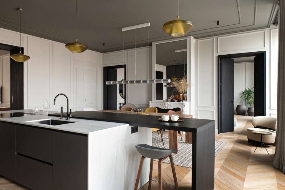 The interior renovation of this home combines the elegance of classical architecture with a contemporary and functional proposal. The open kitchen, featuring dark-toned furniture and marble finishes, becomes the focal point of the space thanks to a fluid layout that visually connects with the dining area and the rest of the rooms. Herringbone flooring, restored woodwork, and decorative lighting reinforce the sophisticated and timeless character of this interior renovation.