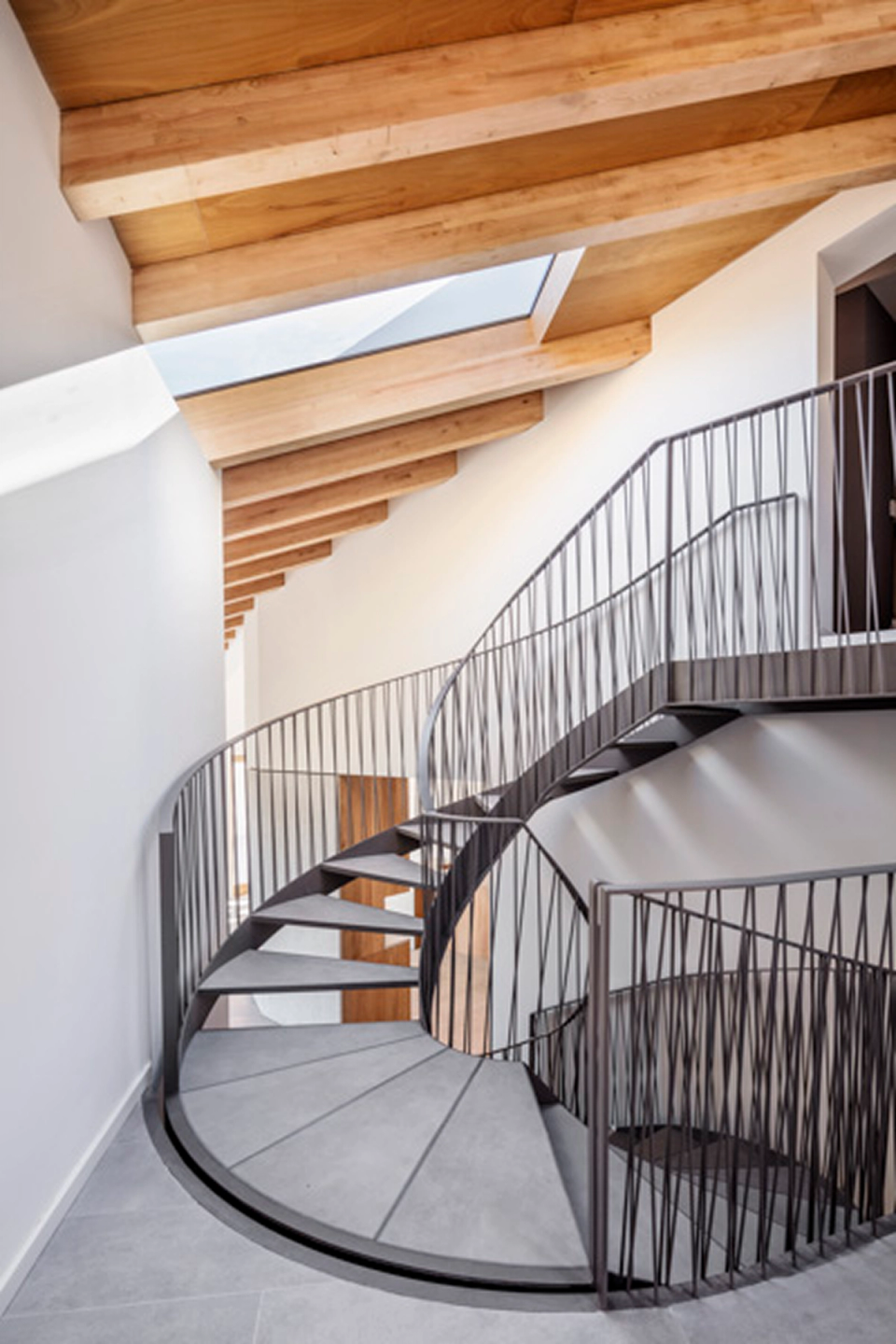 Contemporary helical staircase inside a detached single-family home, featuring a lightweight metal structure, sloped ceilings with exposed wooden beams, and natural light entering through skylights that enhance the sense of spaciousness and warmth of the interior space.