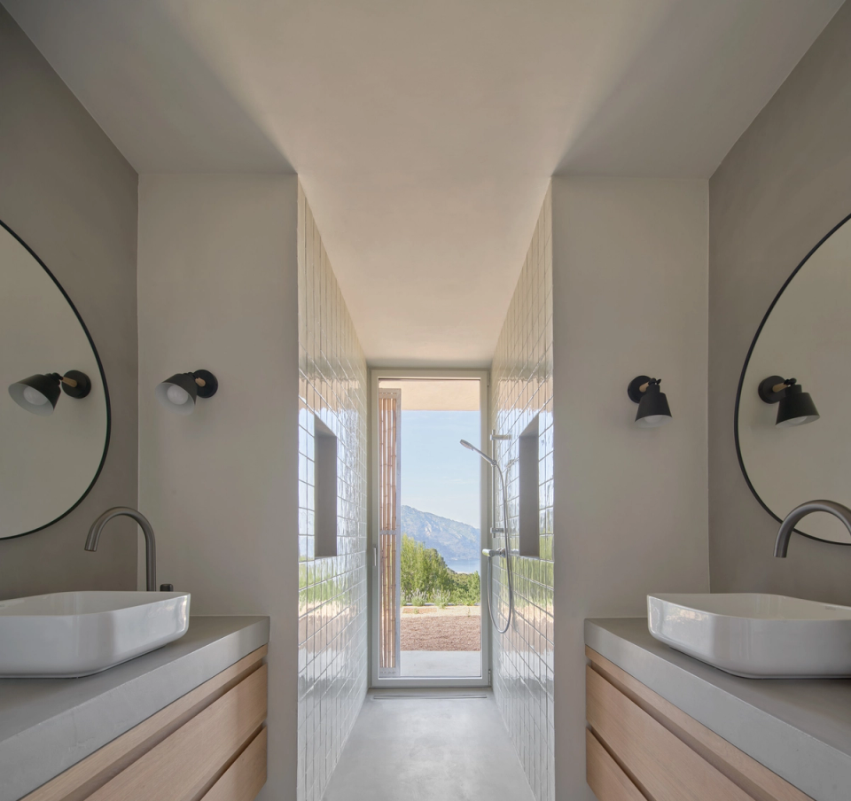 The bathroom of this single-family home stands out for its symmetrical and minimalist layout that enhances natural light and connection with the exterior. Clean lines, light finishes, and wooden furniture create a serene and contemporary atmosphere, while the central opening frames the Mediterranean landscape and adds visual depth to the space. Double sinks, wall lighting, and neutral-toned finishes reinforce the functionality and elegance of the ensemble. This single-family home combines architecture, interior design, and the natural surroundings to create intimate, bright spaces perfectly integrated with the landscape.