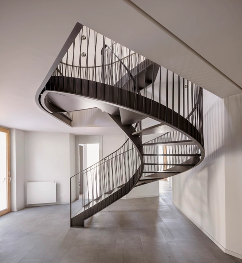 Interior of a contemporary detached single-family home featuring a spectacular organic-shaped metal staircase that visually connects all levels of the house. The space, flooded with natural light through skylights and large openings, combines warm materials such as exposed wooden ceilings with neutral finishes and minimalist lines that reinforce the sense of spaciousness, elegance, and architectural continuity.