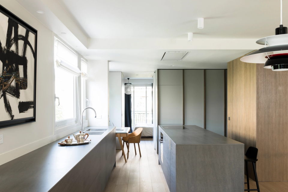 Contemporary-style interior renovation transforming the kitchen into an open, bright, and functional space. The minimalist composition combines gray-toned surfaces with light wood flooring, creating an elegant and balanced contrast. The large central island acts as a gathering point and organizing element of the space, while natural light enhances the warmth of the materials and the sense of spaciousness. The project stands out for its clean lines, integrated furniture, and serene timeless atmosphere.
