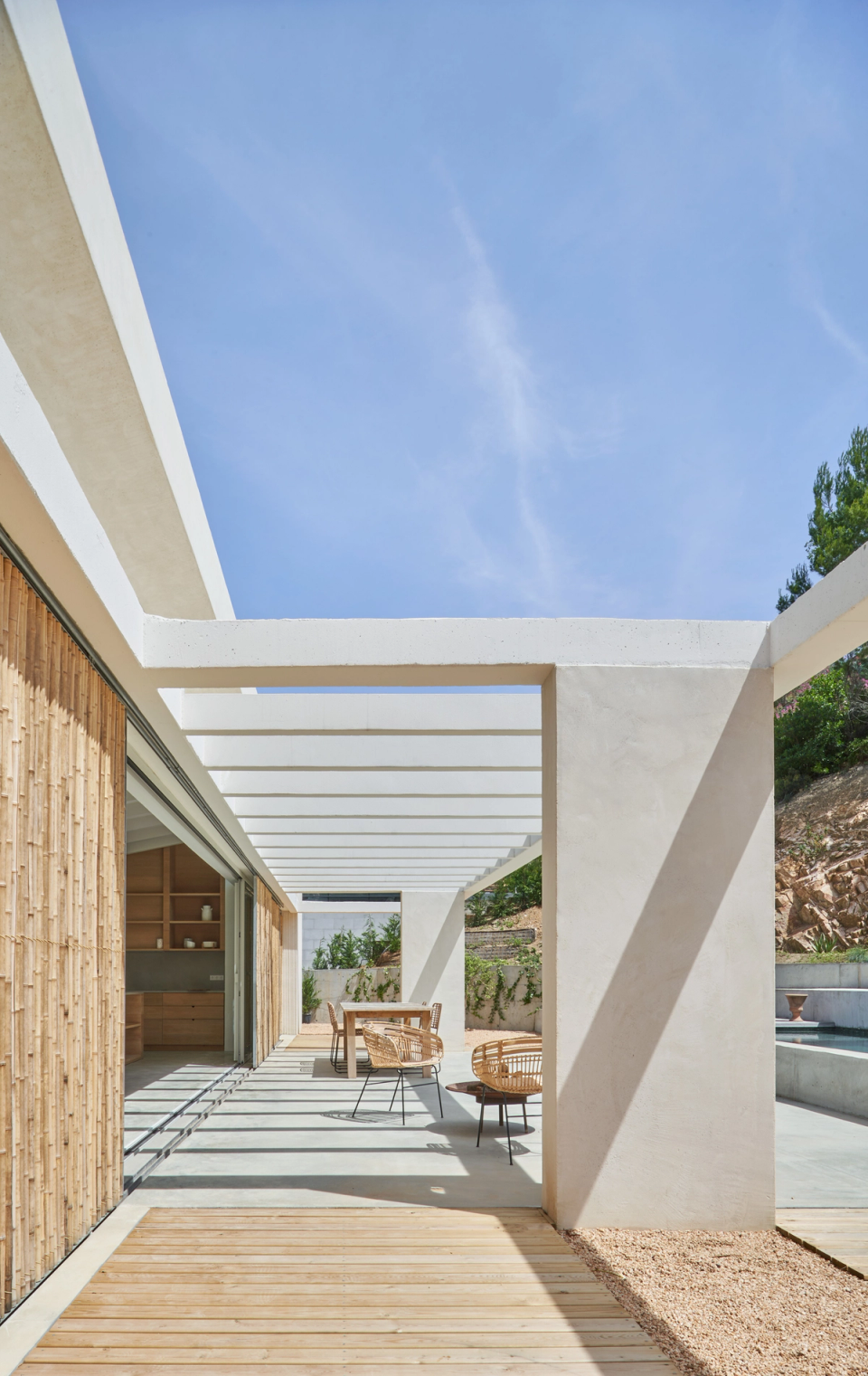 This single-family home embraces light and open exterior architecture, where the structural pergola becomes the main element of the composition. The geometric lines create a play of light and shadow that accompanies the outdoor pathway and reinforces the feeling of depth and spaciousness in the spaces. Natural wood cladding and mineral finishes provide warmth and integrate the home with the surrounding Mediterranean landscape. This single-family home stands out for its fluid relationship between interior and exterior, natural light, and contemporary architecture conceived for enjoying outdoor spaces with comfort and naturalness.