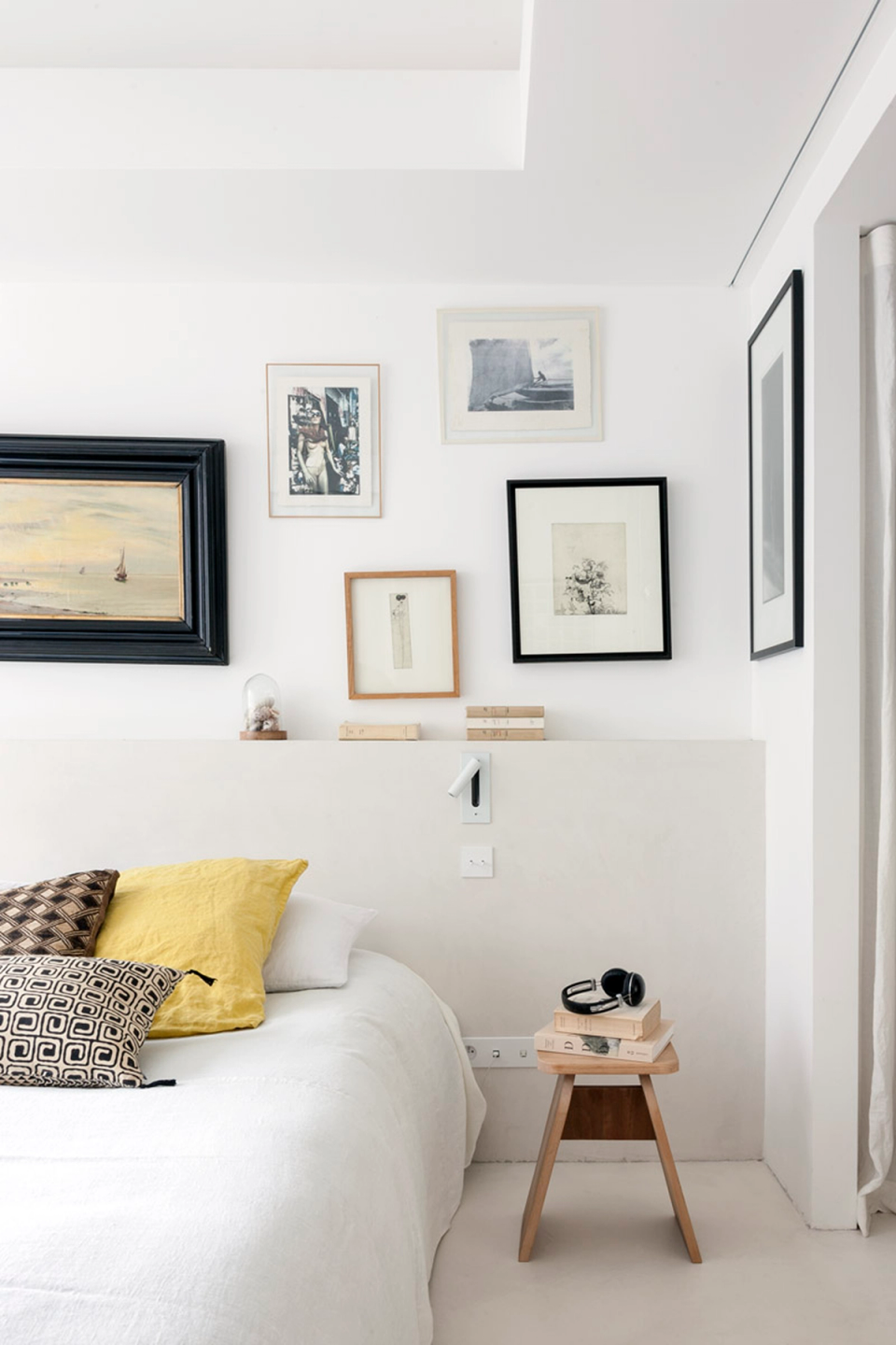 The bedroom interior design combines minimalism, brightness and warmth in a space conceived to convey calm and comfort. The palette of white and neutral tones enhances the sense of spaciousness, while natural textiles and decorative details bring texture and personality. The artistic composition of paintings and decorative pieces above the headboard transforms the wall into a focal point, balancing contemporary elegance and domestic character. Natural wood accents and mustard-colored details reinforce the welcoming atmosphere of this full renovation, where every detail has been carefully designed to create an intimate, functional and harmonious environment.