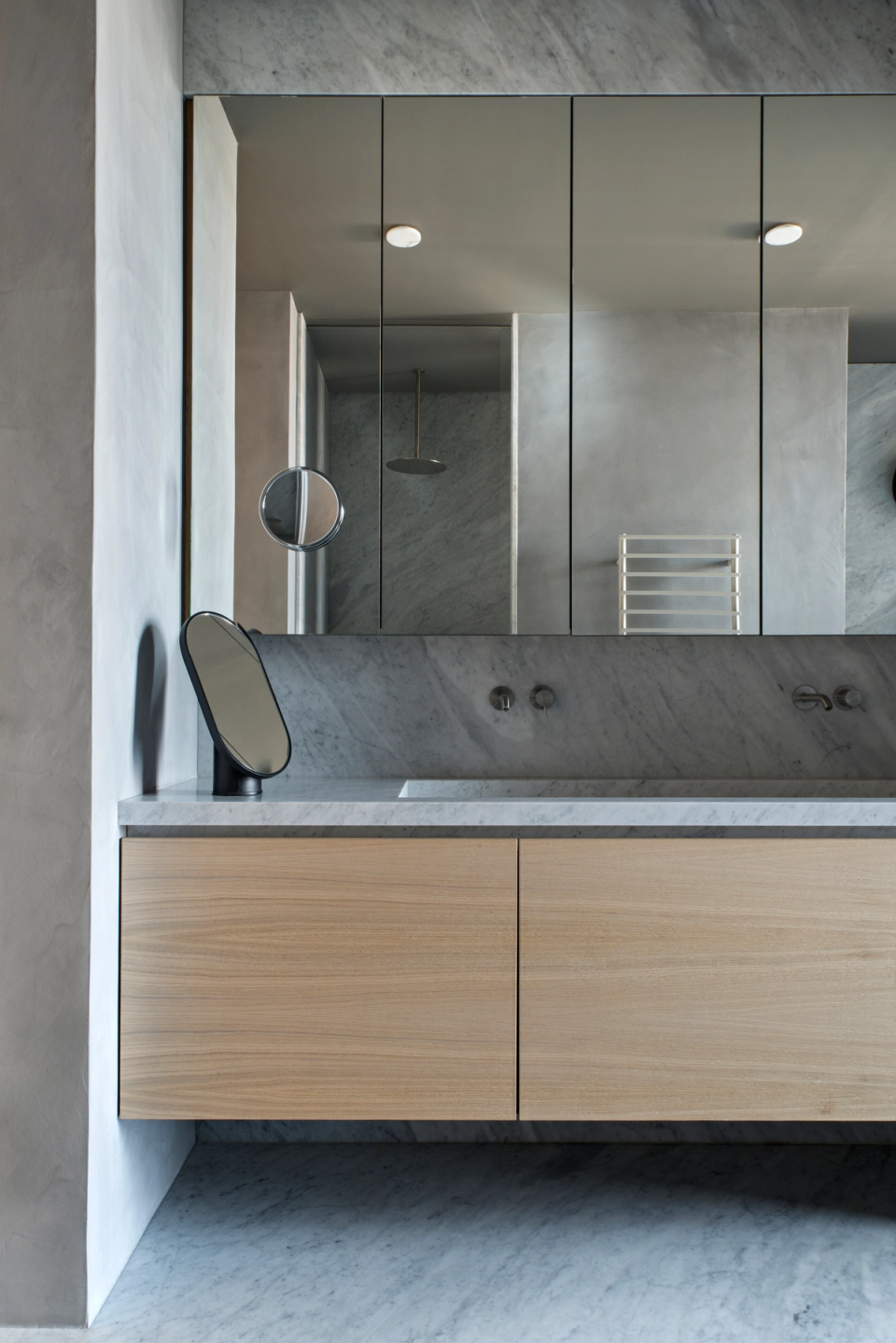 The interior renovation of this bathroom combines minimalism and noble materials to create an elegant, functional, and timeless space. The continuous natural stone cladding provides uniformity and visual depth, while the suspended light-wood furniture introduces warmth and balance to the ensemble. Integrated mirrors, built-in fixtures, and clean lines reinforce a contemporary aesthetic designed to enhance light, order, and the feeling of well-being within the home.