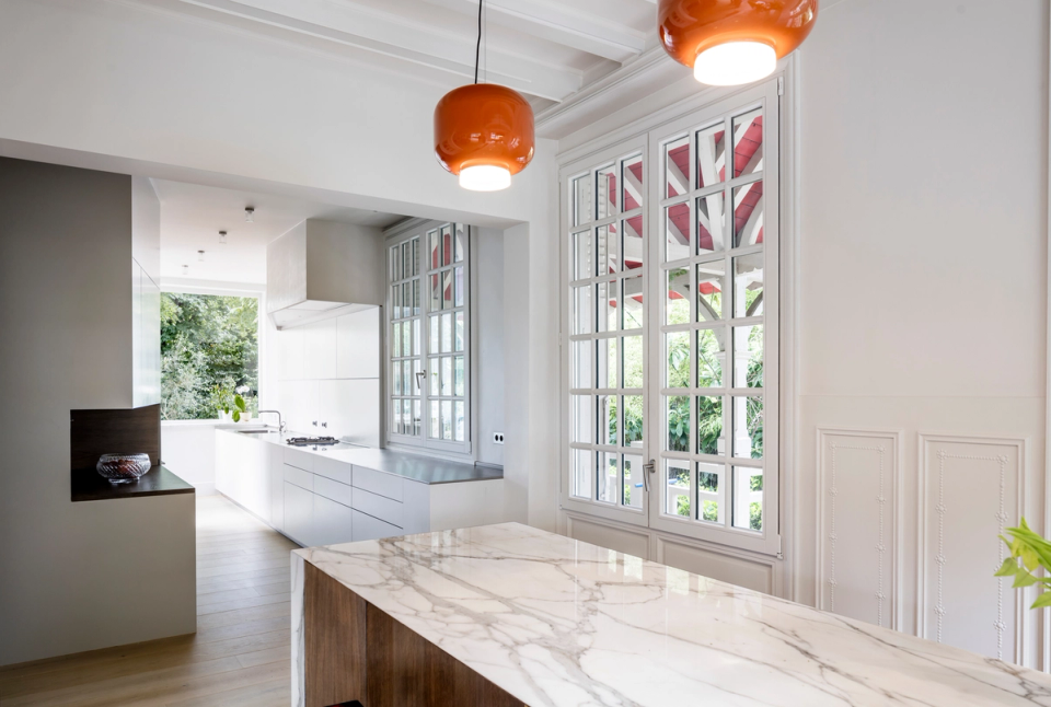 Interior renovation of a connected dining and kitchen space where the original architecture of the residence is highlighted through beam ceilings, molded arches, and large windows. The combination of natural wood furniture, a marble island, and designer pieces in red tones brings warmth, personality, and an elegant contemporary contrast.