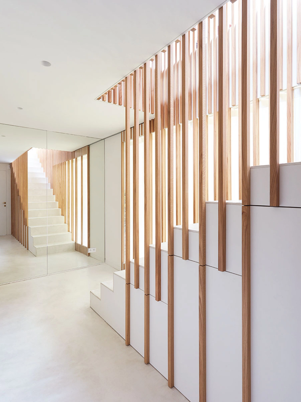 This full renovation enhances natural light and spaciousness through a minimalist architectural proposal with meticulous attention to detail. Vertical wooden slats define the staircase as a sculptural element that organizes the space, filters light and creates an elegant interplay of transparencies and textures throughout the interior.