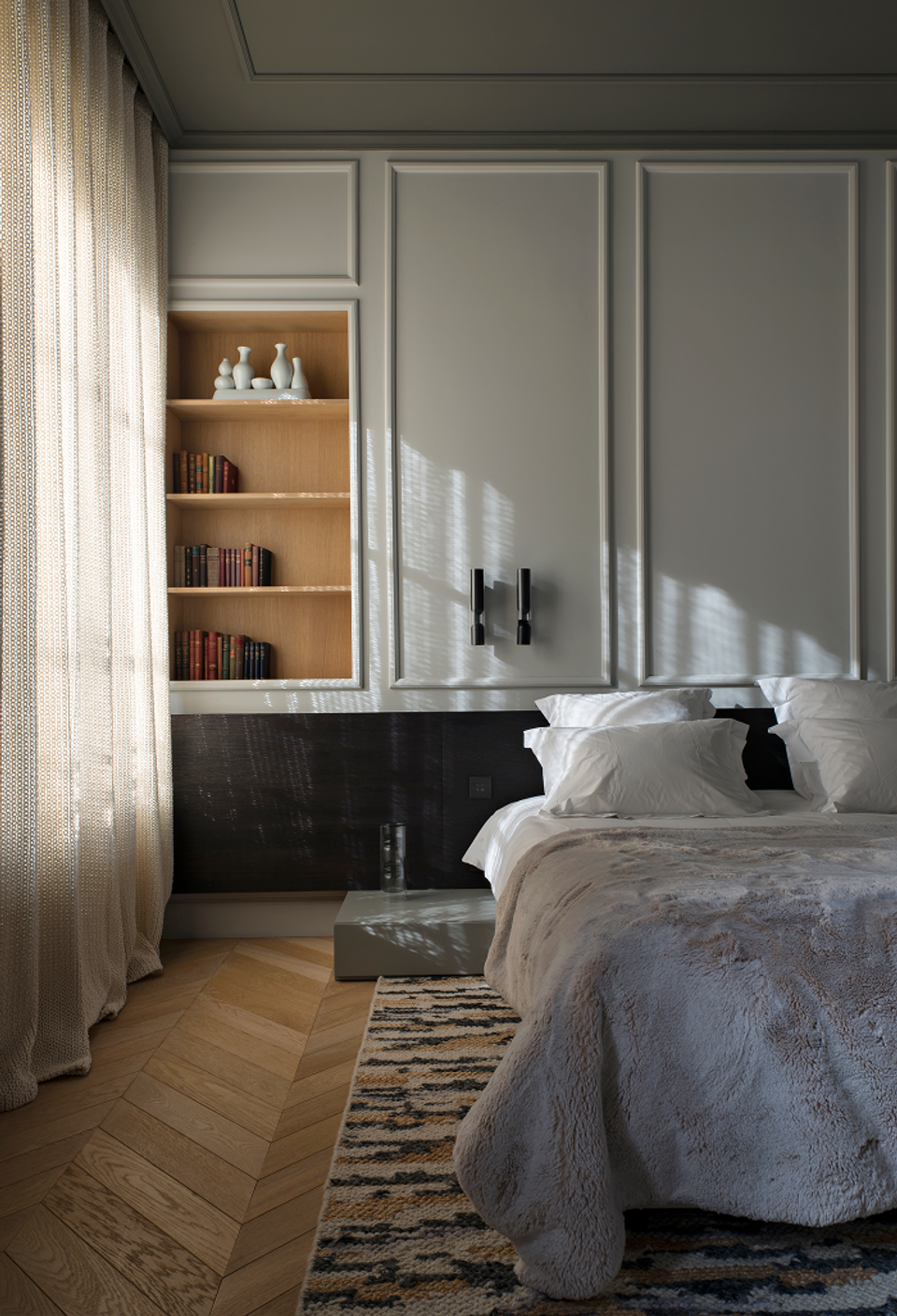 The interior renovation of this bedroom stands out for the balance between sophistication and comfort, combining classical elements with a contemporary and minimalist aesthetic. Molded walls, herringbone parquet flooring, and neutral-toned finishes create a serene and elegant atmosphere, while integrated shelving and subtle lighting provide functionality and visual depth. Natural light filtered through textile curtains reinforces the feeling of intimacy and transforms the space into a warm and welcoming environment designed for relaxation.