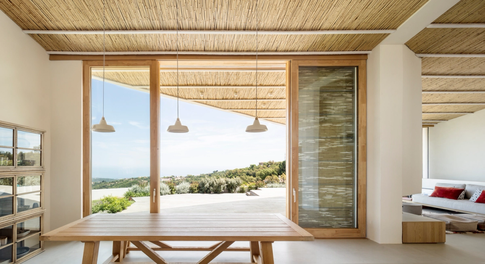 Dining room of a detached house with large panoramic openings framing views of the natural landscape and maximizing natural light inside the home. Ceilings clad with natural materials, wooden furniture and the direct connection with the outdoor terrace create a warm, serene atmosphere perfectly integrated into the Mediterranean surroundings.