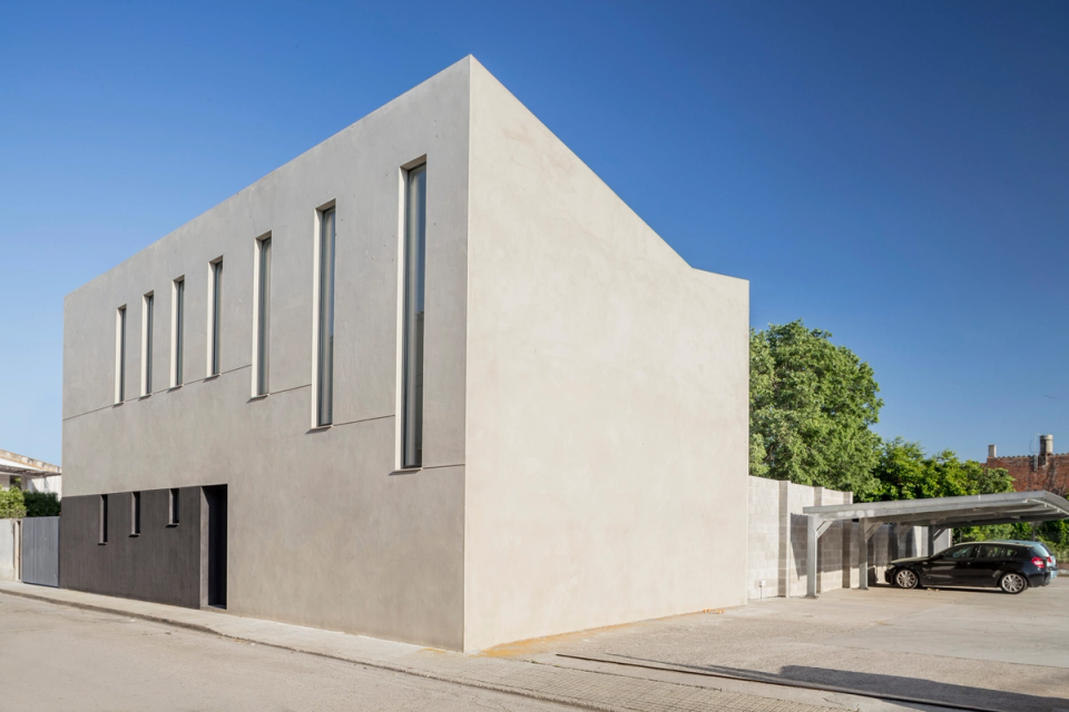 The house appears as a sculptural and silent volume naturally integrated into the urban environment. Continuous surfaces and neutral finishes reinforce the geometric purity of the composition, while the presence of trees and open spaces provides balance, shade, and connection with the outdoor landscape.