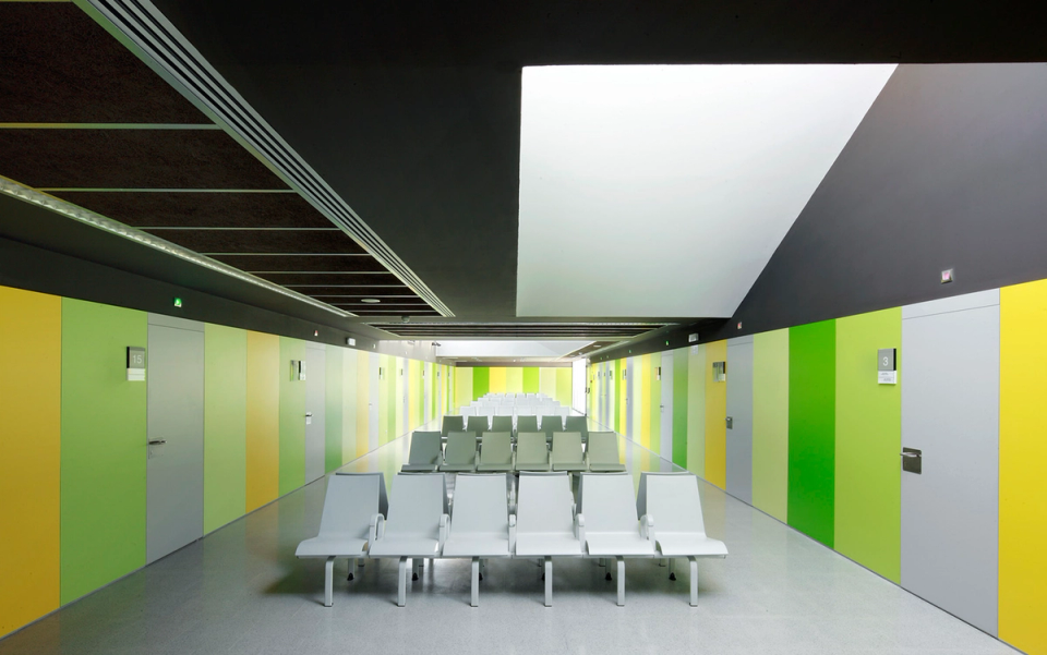 Interior view of the Sant Vicenç Health Center where transparency and connection with the exterior become central elements of the project. Large glass surfaces frame the existing trees and introduce natural light into the common spaces, while the green and yellow color palette provides vitality, visual orientation, and a welcoming atmosphere for the center’s users.