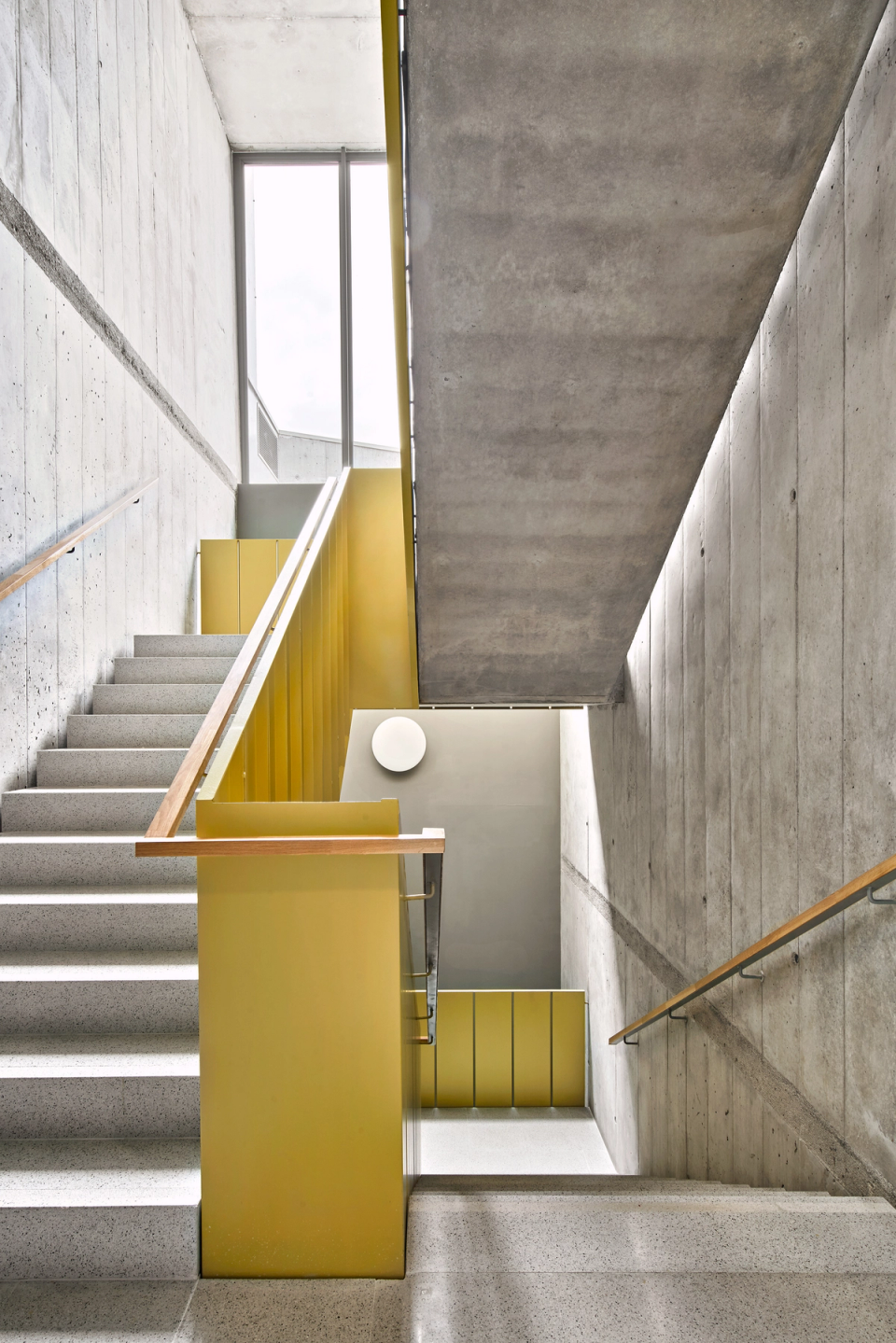 Interior staircase of a contemporary cultural center marked by the contrast between exposed concrete, natural light, and yellow-toned metal cladding that adds identity and warmth to the space. The geometric composition and the entry of light through large windows enhance a functional and elegant architecture designed to connect the different levels of the cultural center with visual spaciousness and fluid circulation.