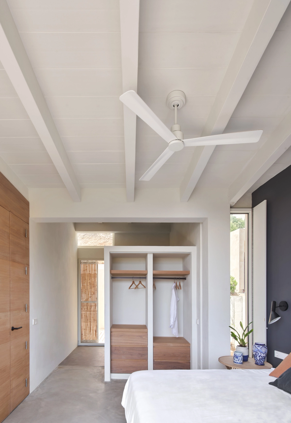 This bedroom in a single-family home embraces essential and luminous architecture, with open spaces and natural materials that reinforce the feeling of comfort and calm. Exposed white beams, wood finishes, and a neutral color palette create a contemporary Mediterranean atmosphere, while vertical openings encourage natural light entry and connection with the exterior. Integrated furniture and minimalist lines maintain a functional and visually clean layout. This single-family home combines design, simplicity, and landscape integration to create warm and timeless residential spaces.