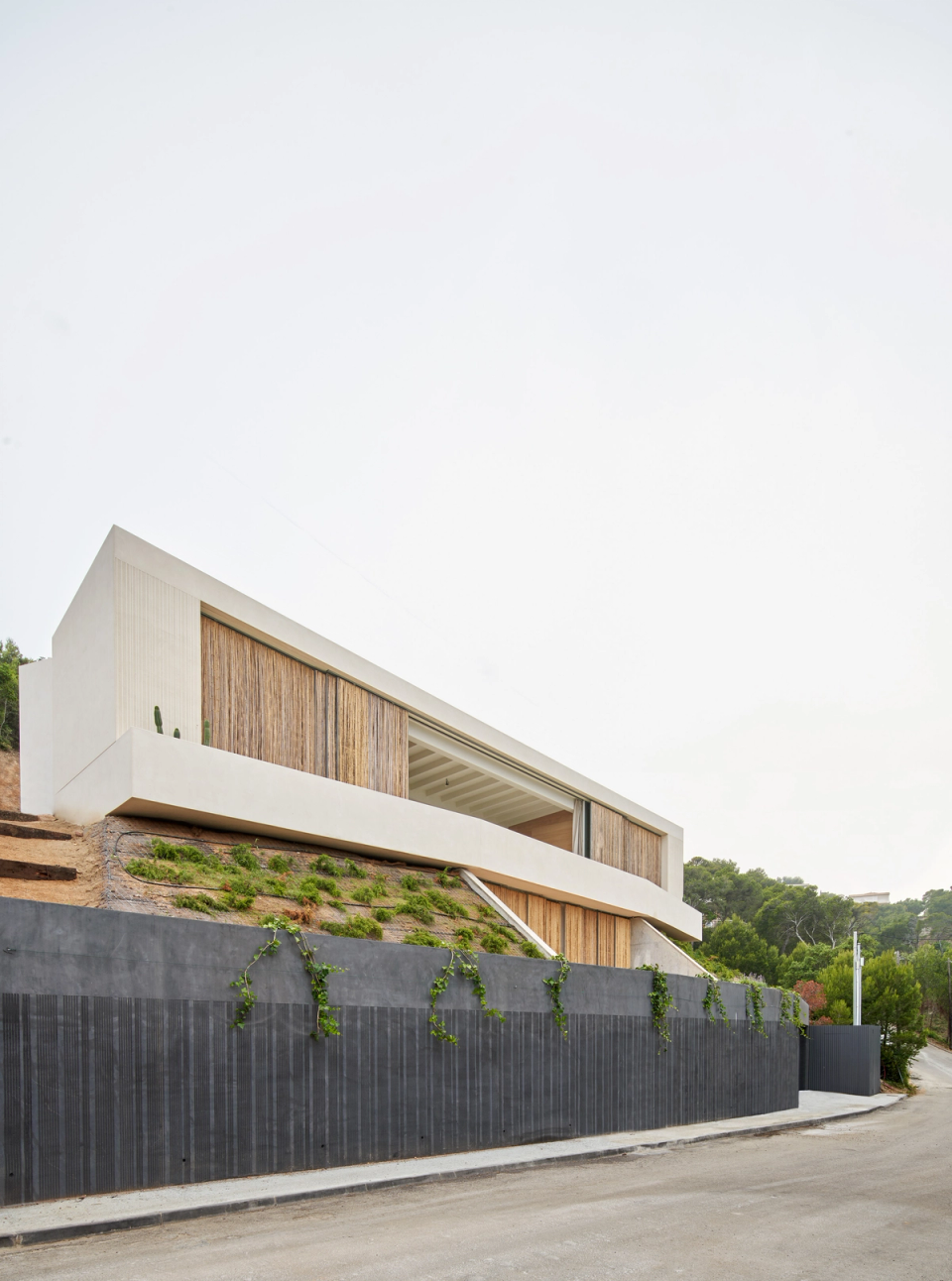 This single-family home features contemporary architecture of great formal purity, defined by linear volumes adapted to the natural slope of the terrain. The project takes advantage of the topography to elevate the main living spaces and enhance views and privacy, while the combination of white surfaces and wooden enclosures provides warmth and integration with the Mediterranean landscape. The terraces and outdoor spaces are naturally articulated with the construction, generating a fluid relationship between interior and surroundings. This single-family home stands out for the balance between minimalism, functionality, and respect for the natural context.