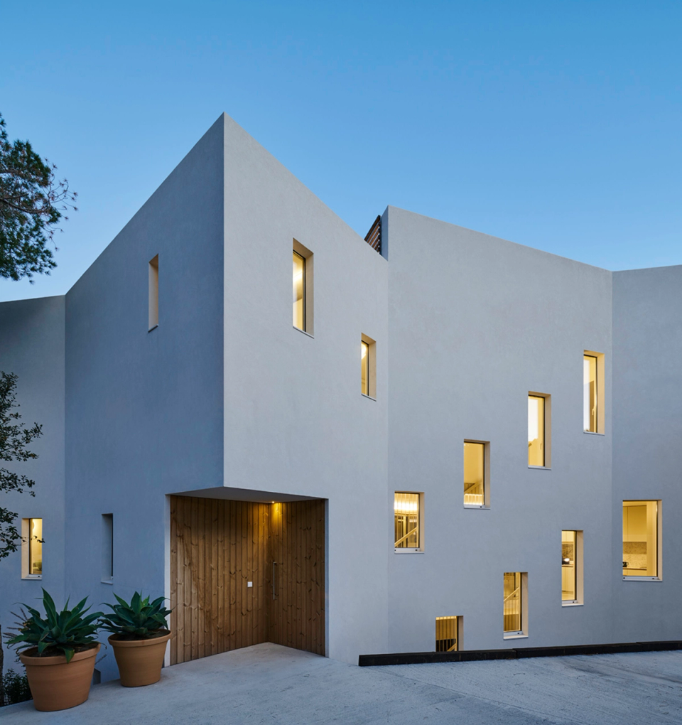 Façade of a contemporary detached single-family home with geometric volumes, white finishes, and vertical openings that bring natural light into the interior spaces. The architecture combines minimalism, natural materials, and integration with the residential environment.
