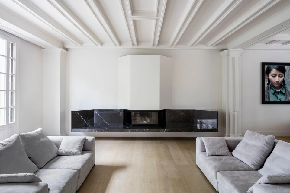 Interior renovation of a living room with a contemporary intervention that respects the classical essence of the residence. The space combines ornate ceilings, original moldings, and a minimalist fireplace with dark stone finishes, creating an elegant, balanced, and naturally bright atmosphere.