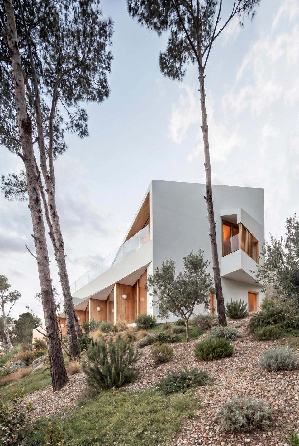 Minimalist detached single-family home located on a steeply sloped plot surrounded by Mediterranean landscape. The project takes advantage of the natural topography to create stepped architecture organically integrated into the terrain, enhancing views and the relationship with the surroundings. White façades, large openings, and wood details provide brightness, warmth, and an elegant contemporary aesthetic. This detached house stands out for its landscape integration, use of native vegetation, and architectural concept designed to connect interior and exterior spaces naturally.