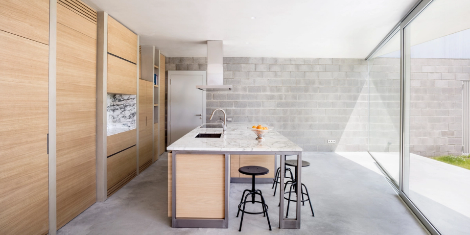 The kitchen is designed as an open, restrained, and functional space where natural light becomes the protagonist thanks to large glazed openings directly connected to the outdoor courtyard. The white marble island organizes the space and adds elegance, while the light wood furniture and metal profiles define a warm contemporary aesthetic. Exposed block walls and continuous concrete flooring reinforce the honest and minimalist character of the house, creating a serene and balanced atmosphere.