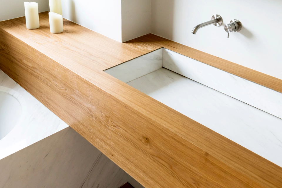 Interior reform of a contemporary minimalist bathroom with integrated toilet, natural wood baking plate and stainless steel wall taps. The space stands out for its clean lines, natural light and the harmonious combination of white tones and warm wood finishes, creating an elegant, serene and functional atmosphere. Modern interior design with refined aesthetics, noble materials and sophisticated design oriented to well-being and calm