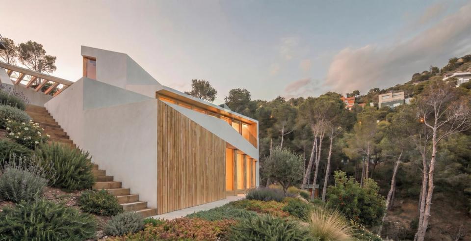 Contemporary detached single-family home integrated into a natural setting of great landscape value, designed to adapt sensitively to the sloping plot. The project combines minimalist architecture, white façades, and vertical wood cladding that brings warmth and reinforces the connection with nature. Large openings allow natural light to enter and establish a continuous relationship between interior spaces and the surrounding landscape. This detached house stands out for its organic integration into the terrain, Mediterranean vegetation, and architecture focused on views, privacy, and comfort.
