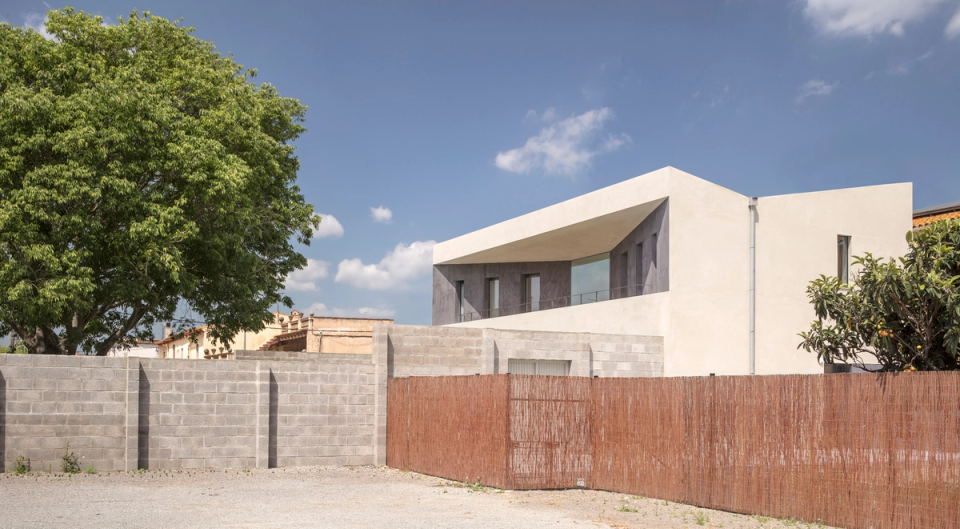 The exterior volumetry stands out for its pure and minimalist geometry, defined by continuous light-toned planes and narrow vertical openings that organize the façade with rhythm and precision. The house is presented as a compact and monolithic piece contrasting with the lightness of the lateral open spaces and the transparency of the interior. The austere composition, together with natural light and neutral materials, reinforces the contemporary and serene character of the architecture.