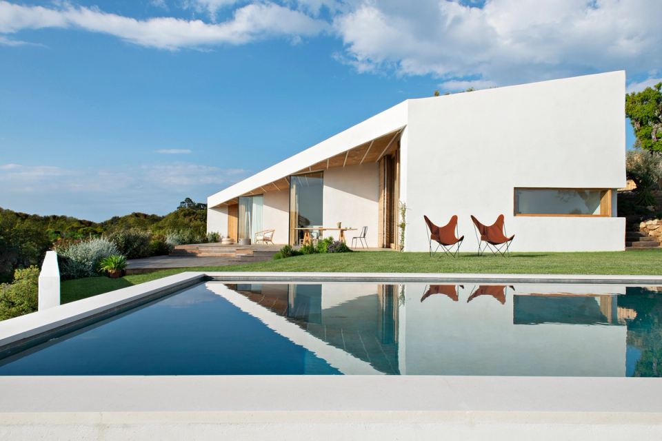 Outdoor area of a detached house with a swimming pool integrated into a natural environment and open views towards the landscape. The combination of Mediterranean vegetation, wooden terrace and relaxation areas creates a calm and bright atmosphere designed to strengthen the connection between architecture, nature and outdoor living.