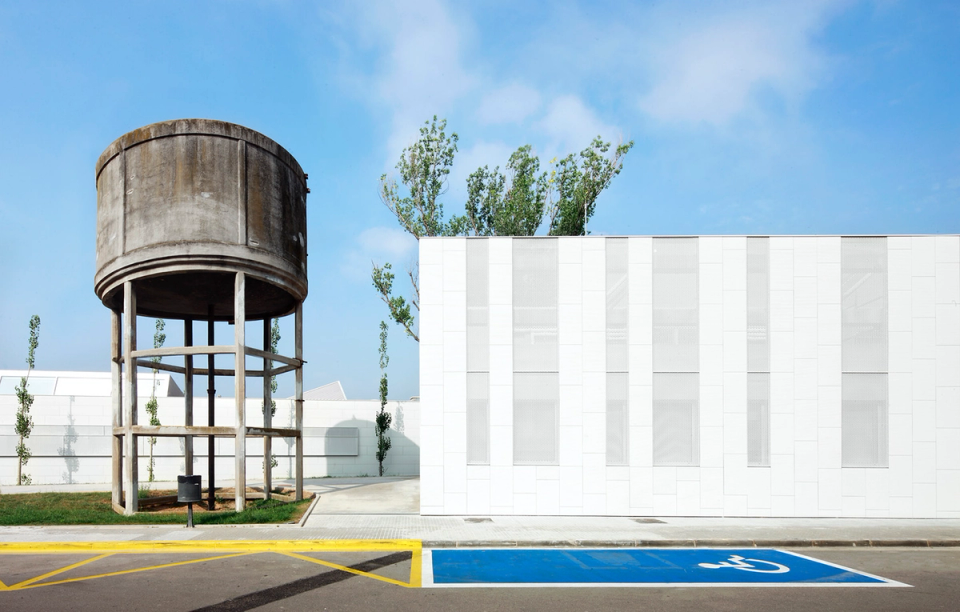 Exterior detail of the Sant Vicenç Health Center where contemporary architecture and industrial heritage coexist. The project integrates the existing water tower as an emblematic and historical memory element, contrasting with the new white façade featuring vertical lines and minimalist finishes. The clean composition, natural light, and open spaces reinforce the modern and functional identity of this healthcare facility.
