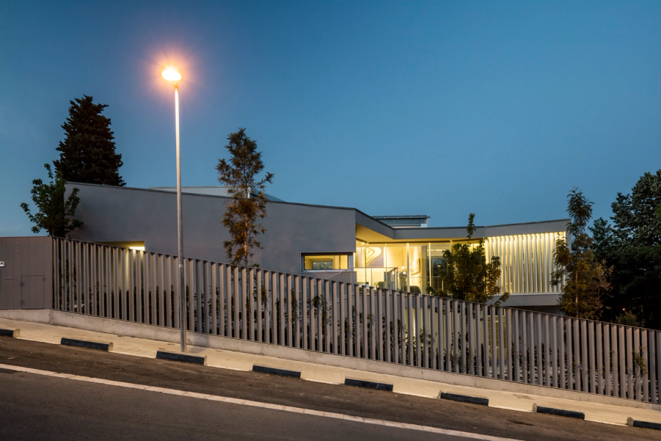 Night view of a contemporary single-family home with elegant and minimalist architecture highlighted by integrated lighting and the relationship between interior and exterior spaces. The horizontal volumes, large glass surfaces, and vertical slats create a sophisticated architectural composition that becomes especially prominent at dusk thanks to the warm interior lighting. The house adapts to the slope of the terrain with a discreet and integrated layout, while the linear enclosure and neutral-toned finishes reinforce the modern aesthetic of the ensemble. A single-family residential project that combines design, functionality, and landscape integration with a contemporary and timeless image.