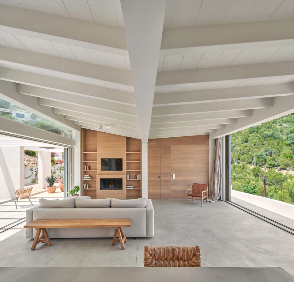 The interior of this single-family home is defined by open, bright architecture fully connected to the surrounding natural landscape. Large panoramic openings integrate the views into the living spaces, while the sloped roof with exposed white wooden structure provides spaciousness, rhythm, and Mediterranean character. Warm natural wood finishes and minimalist furniture create a serene and elegant atmosphere where natural light becomes the protagonist. This single-family home combines functionality, comfort, and contemporary design to create spacious areas designed to enjoy the connection between interior and exterior.