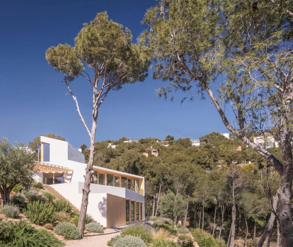 Detached single-family home with minimalist lines and compact volumetry, conceived to blend naturally into a mountain setting with open views of the Costa Brava landscape. The combination of white façades and wood cladding adds warmth and reinforces the contemporary character of the project. Vertical openings filter natural light and create a subtle relationship between interior and exterior spaces while maintaining privacy. This detached house stands out for its refined architecture, adaptation to the site’s topography, and placement designed to maximize views and connection with the natural environment.