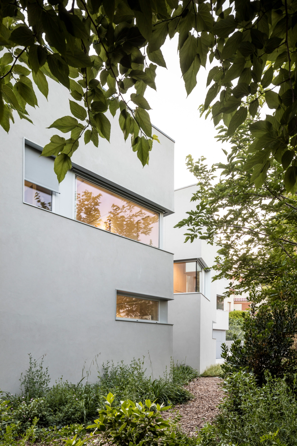 Exterior view of a contemporary single-family home integrated into a natural landscaped environment, featuring clean lines and geometric volumes that prioritize light, privacy, and the relationship with the outdoors. The soft-toned façades combine with large horizontal openings that frame the views and allow a constant visual connection with the surrounding vegetation. The outdoor pathway among trees and plant species reinforces the sense of calm and intimacy, while the minimalist composition of the house provides an elegant, modern image perfectly integrated into the residential landscape.