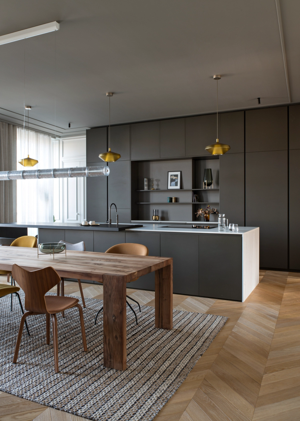 The interior renovation of this kitchen embraces an elegant and contemporary aesthetic, where the dark finishes of the furniture contrast with the warmth of the herringbone parquet flooring and the natural wood of the dining area. The new open layout enhances functionality and the connection between spaces, while decorative lighting and noble materials add character, sophistication, and a welcoming atmosphere.