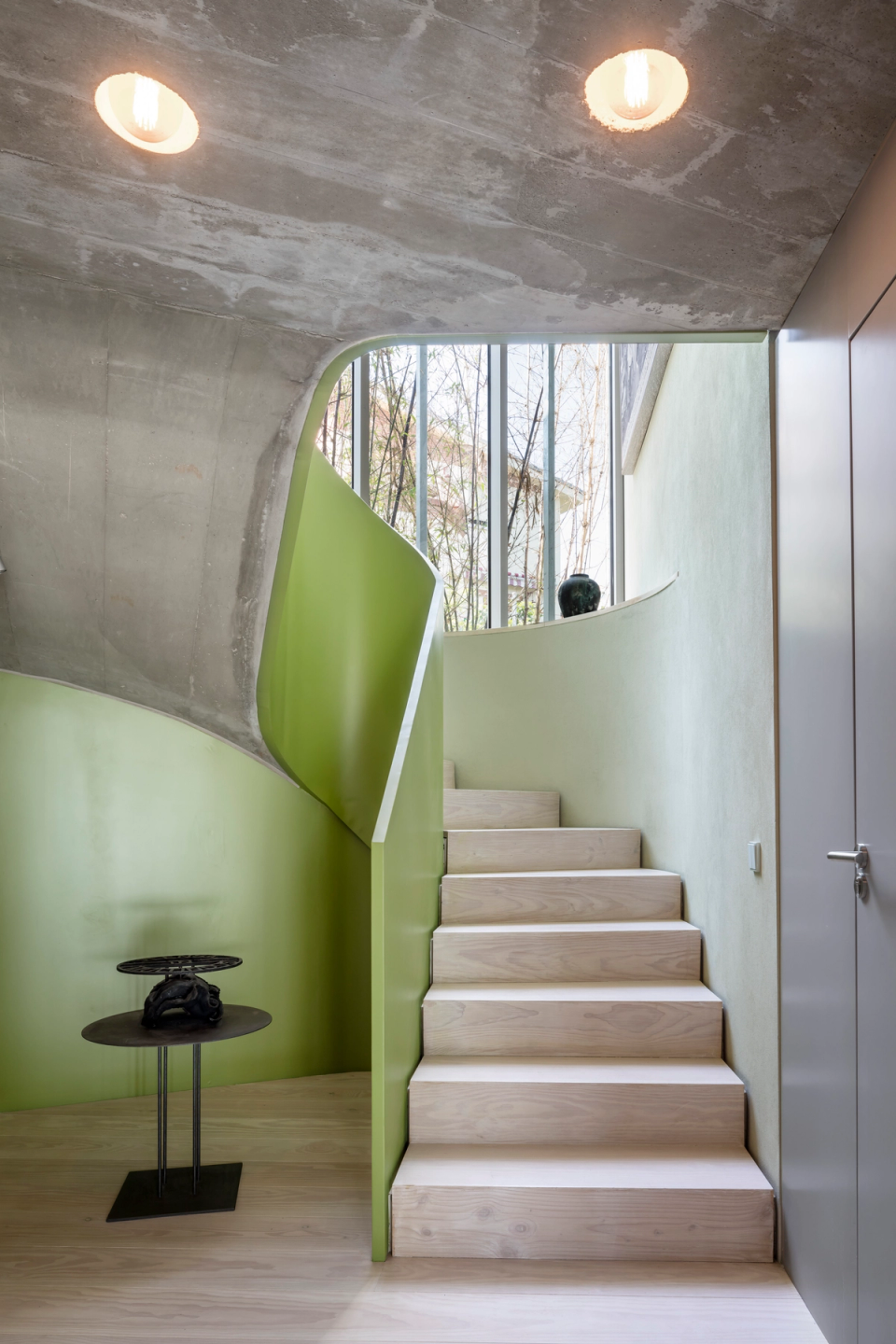 Interior staircase of a contemporary single-family home with a sculptural composition that combines organic forms, exposed concrete, and colorful finishes. The curved green-toned railing brings dynamism and personality to the space, while the natural light entering through the vertical window reinforces the feeling of spaciousness and connection with the outdoors. The combination of materials such as light wood, concrete, and smooth surfaces creates a modern, elegant, and architecturally distinctive atmosphere.