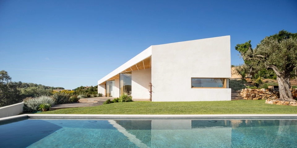 Exterior of a minimalist detached house with clean volumes and pure geometries harmoniously integrated into the natural landscape. White façades, large glass surfaces and a pitched roof with wooden finishes bring brightness and warmth to the home, while the swimming pool and landscaped areas strengthen the connection between architecture, outdoor living and the Mediterranean lifestyle.