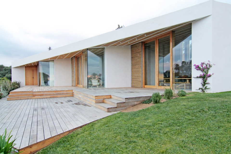 Exterior of a modern detached house with an elegant architectural composition based on horizontal lines, large glass surfaces and natural wood finishes. The elevated terrace and landscaped areas create a fluid transition between interior and exterior, while the pitched roof protects the open spaces and reinforces the contemporary Mediterranean character of the home.