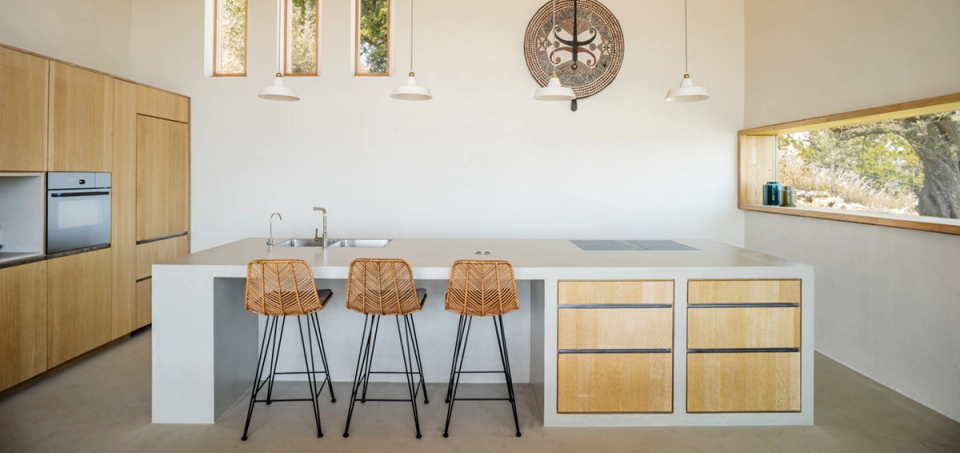 Contemporary kitchen integrated into a detached house, featuring a large central island, natural wood cabinetry and an open layout that enhances functionality and brightness. Vertical openings and the panoramic window provide natural light and connection with the outdoor environment, while minimalist lines and warm materials reinforce the elegant Mediterranean aesthetic of the home.