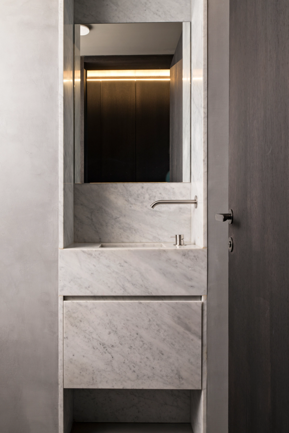 The interior renovation of this bathroom embraces a minimalist and refined aesthetic, where natural marble becomes the main protagonist of the space. The integrated vanity, clean lines, and built-in fixtures provide a sense of order and sophistication, while indirect lighting enhances the depth and warm atmosphere of the overall design. The combination of neutral tones and dark finishes creates an elegant and contemporary contrast that transforms this space into a functional area of high aesthetic value.