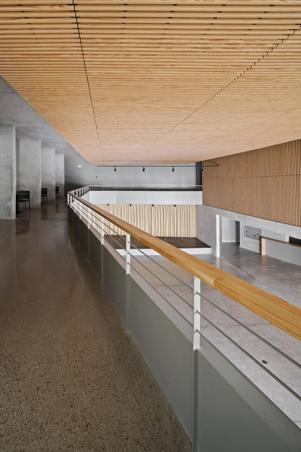 Interior detail of a cultural center where the wooden slat ceiling becomes the main feature of the architectural space. The linear geometry and natural finish provide acoustic comfort, warmth, and a contemporary identity to this multipurpose area of the cultural center. The combination of exposed concrete, large openings, and integrated furniture creates a functional and elegant atmosphere designed to host cultural, social, and institutional activities.