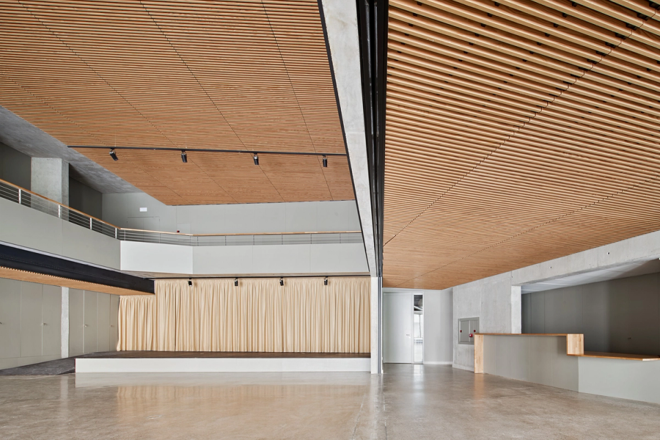 Large multipurpose hall in a cultural center designed to provide flexibility, spaciousness, and comfort for public activities and cultural events. The space stands out for its continuous wooden slat ceilings, which provide acoustic quality and a warm atmosphere, combined with exposed concrete structures and large openings visually connected to the exterior. The open layout and natural light reinforce the contemporary character of the cultural center, prepared to host exhibitions, conferences, social events, and community activities.