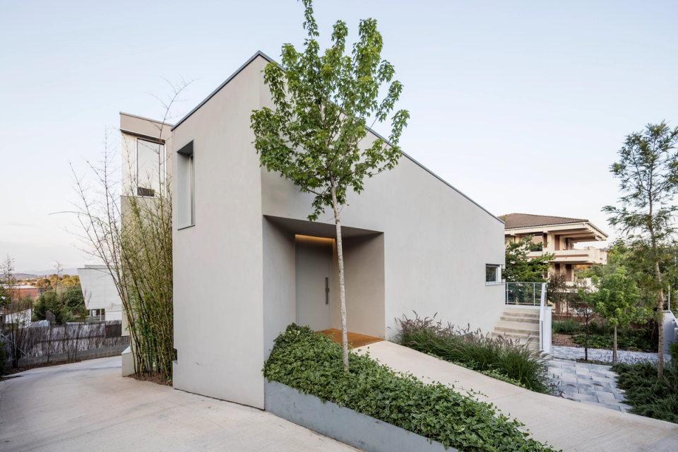 Main entrance of a contemporary single-family home with minimalist architecture defined by pure volumes, inclined lines, and a sober yet elegant aesthetic. The light-toned façade combines with integrated vegetation and continuous paving that reinforce the relationship between architecture and landscape. The entrance is framed within a recessed volume that provides depth and protection, while vertical and horizontal openings allow natural light to enter while maintaining the privacy of the interior spaces. A modern single-family home project that stands out for its formal simplicity, integration with the surroundings, and balance between functionality and architectural design.