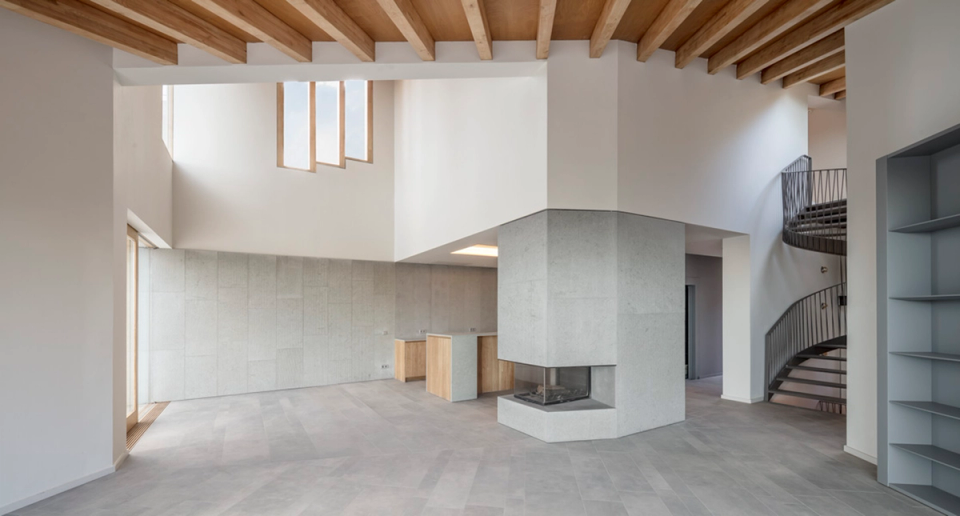 Large open-plan space in a contemporary detached single-family home integrating living room, kitchen, and circulation areas beneath exposed wooden ceilings that provide warmth and visual continuity. The central fireplace acts as the organizing element of the house, while the curved metal staircase and vertical openings reinforce the sculptural character and natural brightness of the architectural project.