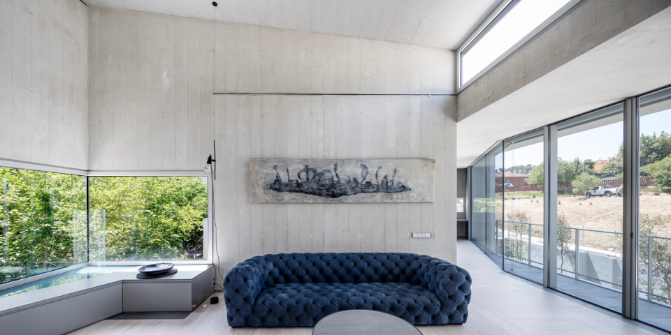Living room of a contemporary single-family home with open-plan spaces, large panoramic windows, and exposed concrete finishes that provide a modern and highly refined architectural aesthetic. The double-height space and floor-to-ceiling openings enhance natural light and the visual connection with the outdoors, while designer furniture and artistic pieces create a sophisticated, elegant, and personality-filled atmosphere.