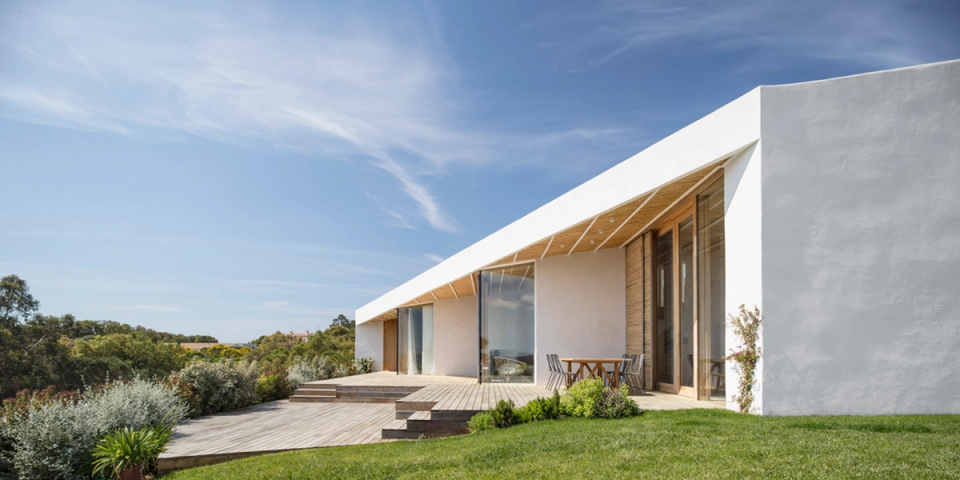 Outdoor terrace of a contemporary detached house with clean architectural lines and a pitched roof that creates protected spaces open to the landscape. White façades, natural wood enclosures and large glass surfaces enhance the connection between interior and exterior, creating a bright, elegant atmosphere fully integrated with the Mediterranean surroundings.
