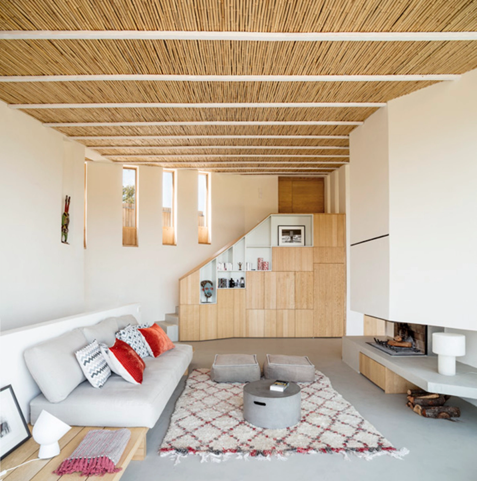 Living room of a detached house with a warm Mediterranean atmosphere defined by ceilings clad with natural materials, large glass enclosures and sliding natural fiber screens that regulate natural light. The interior opens completely towards the terrace and outdoor landscape, creating a fluid connection between architecture, nature and contemporary comfort.