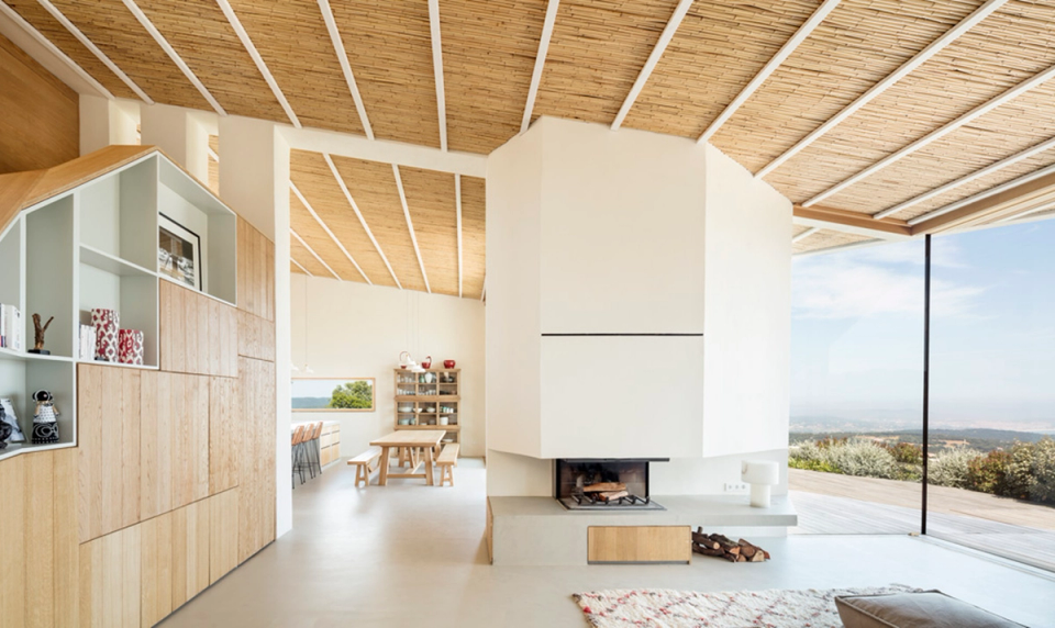 Interior of a detached house with open spaces and warm architecture defined by ceilings clad in natural wood and abundant natural light. The central fireplace acts as the focal point of the living area, visually connecting the living room, dining room and outdoor terrace, while large panoramic openings strengthen the relationship between the home and the landscape.