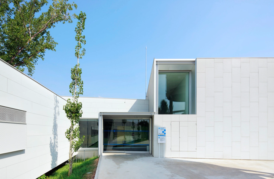 Exterior view of the Sant Vicenç Health Center highlighting the relationship between the new healthcare architecture and the existing heritage. The project combines white volumes with refined lines and the preservation of the original water tower, transformed into a singular feature that provides identity and historical memory to the complex. The integration of vegetation, natural light, and open spaces reinforces the welcoming, functional, and contemporary character of the facility.
