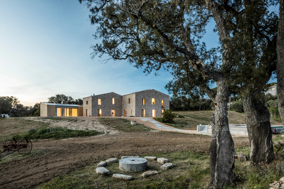 Exterior façade of a comprehensive renovation that combines traditional stone architecture with contemporary volumes featuring pure lines and large glazed surfaces. The project restores the original essence of the rural construction while incorporating new open, bright spaces connected to the outdoors. The composition of natural materials, warm lighting, and the minimalist design of the exterior paving reinforce the elegant and timeless character of this full renovation, conceived to integrate comfort, landscape, and modern architecture into a harmonious whole.