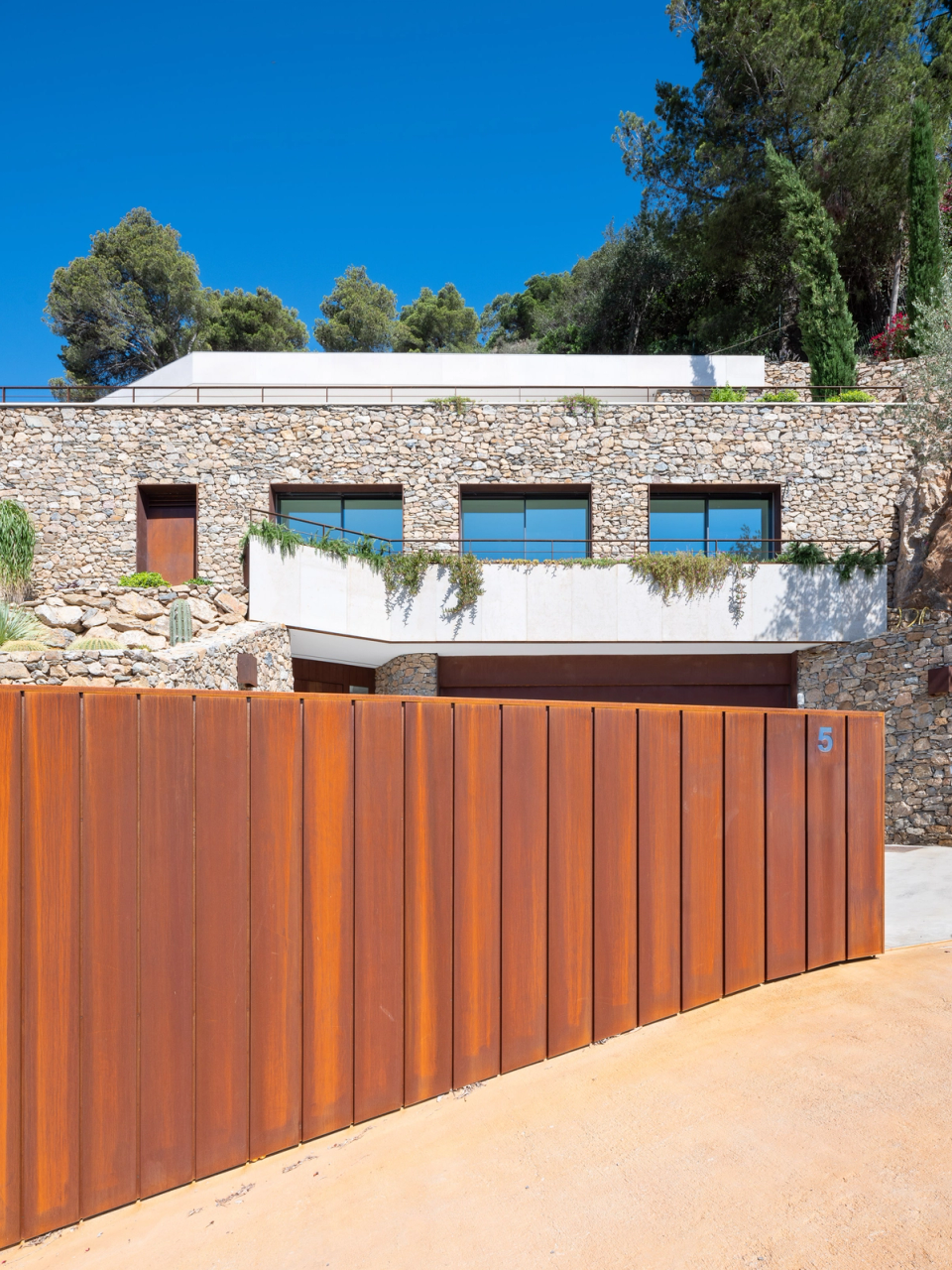 Contemporary detached single-family home combining pure lines, natural materials, and respectful integration with the surroundings. The façade alternates natural stone, wood cladding, and large glazed surfaces that provide brightness and create a constant connection between interior spaces and the outdoors. This architectural and interior renovation embraces a minimalist and warm aesthetic, where every detail is designed to enhance functionality, comfort, and the relationship with the Mediterranean landscape.